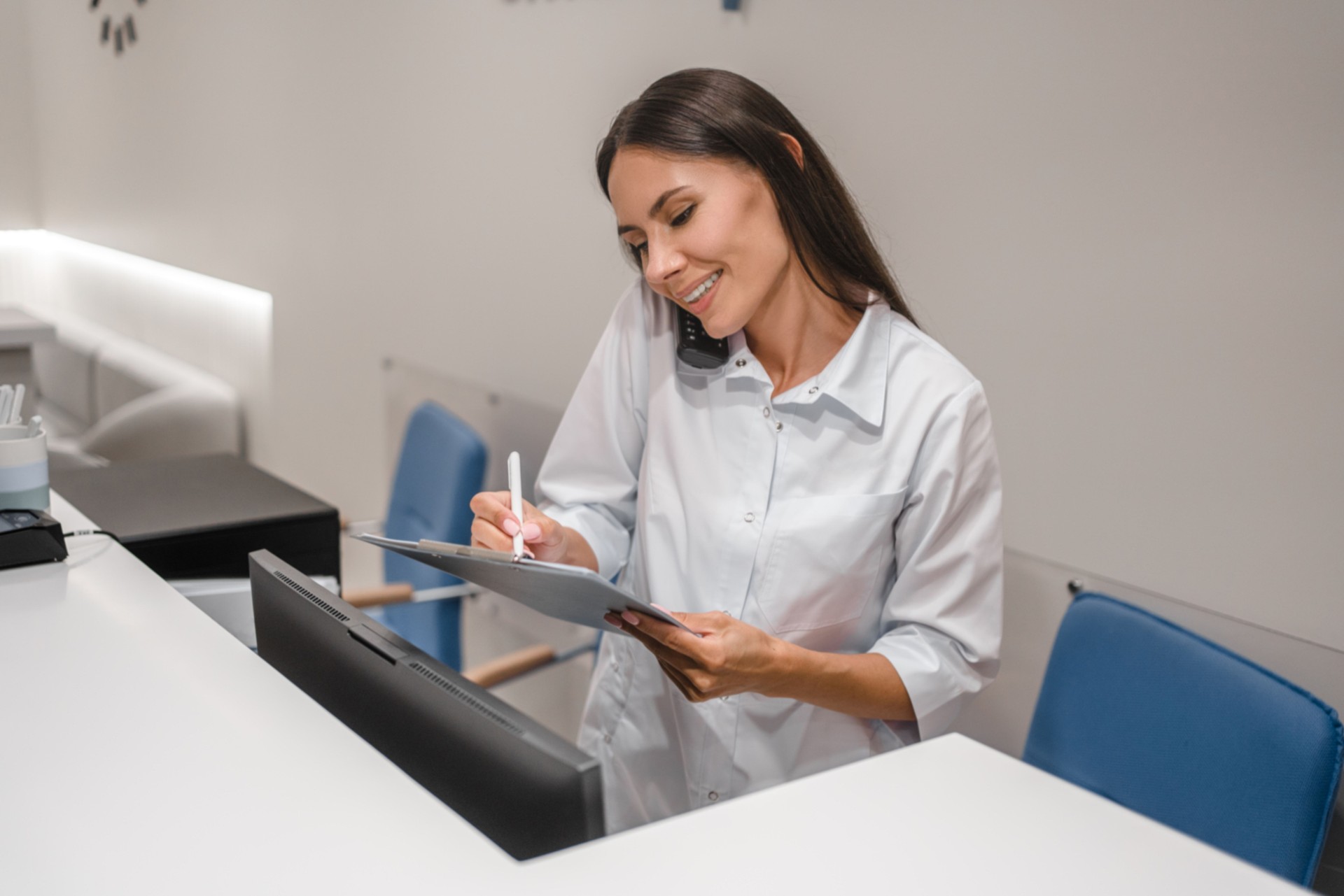 Hospital front desk female receptionist practitioner nurse talking on the phone with customers patients and making doctor appointment and writing on clipboard in hospital.